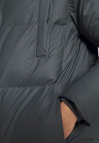 Chaqueta acolchada en gris oscuro, con un bolsillo en el pecho con cierre, puños elásticos y una textura suave, diseñada para el aislamiento y la comodidad.