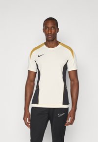 Camiseta de manga corta de Nike en crema con detalles en negro y dorado; presenta un diseño texturizado, cuello redondo y silueta ajustada.
