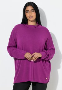 Frau mit langen schwarzen Haaren, die ein locker sitzendes, langärmeliges lila Oberteil und schwarze Hose trägt, steht vor einem weißen Hintergrund.