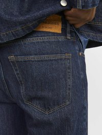 Jeans din denim de culoare bleu închis au un design standard cu cinci buzunare, accentuat cu cusături portocalii și o etichetă din piele maro a brandului pe spate.