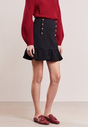 Femme portant une mini-jupe noire à volants avec des boutons dorés, un pull en maille rouge et des mocassins rouges à enfiler avec des accents de boucle dorée.