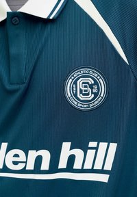 Maillot de sport bleu sarcelle foncé avec col blanc, écusson circulaire du Athletic Club et texte blanc "den hill" sur la poitrine.