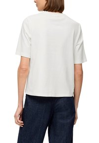 Biała koszulka z krótkim rękawem, okrągłym dekoltem, wykonana z miękkiej bawełny, o luźnym kroju, widoczna z tyłu, w zestawieniu z ciemnymi dżinsami.