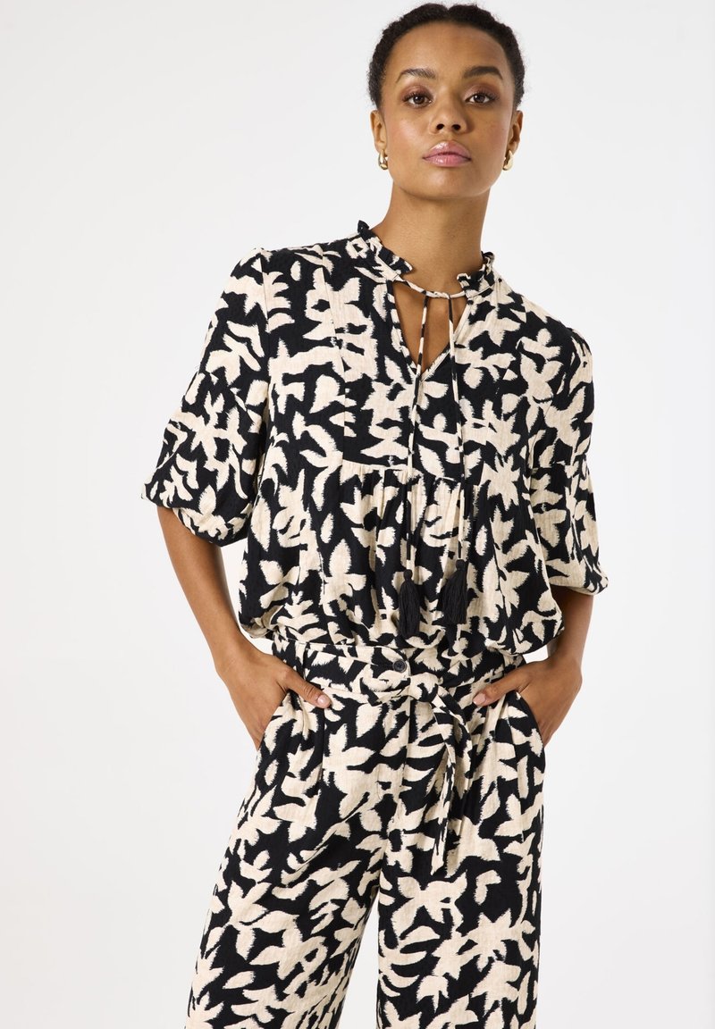 Schwarz-weiße gemusterte Bluse mit gebundenem Kragen, kurzen Ärmeln und lockerer Passform, kombiniert mit passenden weit geschnittenen Hosen.