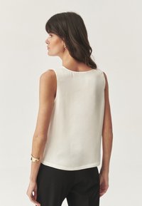 Chemisier blanc sans manches avec une texture lisse, un col rond et une coupe légèrement ample, associé à un pantalon noir et un bracelet à accent doré.