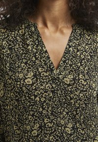 Blouse noire avec un col en V, présentant un motif floral jaune et abstrait. Fabriquée en tissu léger avec une finition texturée.