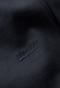Tela de algodón negra con un logo bordado de "Superdry", que presenta una textura suave y un mínimo detalle de costura.