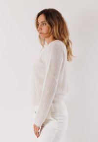 Donna con lunghi capelli mossi che indossa un maglione bianco a maglia e pantaloni bianchi, in piedi di profilo contro uno sfondo chiaro e semplice.