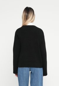 Svart stickad tröja med avslappnad passform, långa ärmar och ribbat avslut. Bärs över en vit skjorta med krage, tillsammans med ljusblå jeans.