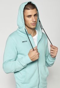 Sudadera de cremallera azul claro hecha de tela suave, con una gran capucha, cordones negros, bolsillos laterales y un pequeño logo en el pecho.