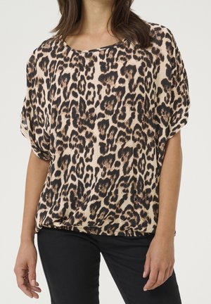 Kobieta ubrana w luźną bluzkę z krótkim rękawem w brązowo-czarny wzór w panterkę, zestawioną z czarnymi spodniami.