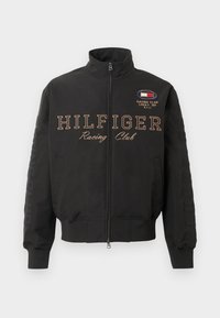 Jachetă neagră cu fermoar, cu manșete ribbate, textul "HILFIGER Racing Club" pe față și un mic emblema de curse pe pieptul din dreapta sus.