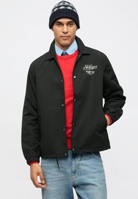 Μαύρο ελαφρύ μπουφάν με κουμπώματα, με κεντημένο λογότυπο, φορεμένο πάνω από ένα κόκκινο πουλόβερ και ανοιχτό μπλε πουκάμισο, συνδυασμένο με τζην παντελόνι.