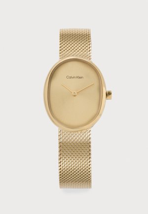 Montre-bracelet Calvin Klein dorée avec cadran ovale, cadran minimaliste montrant les aiguilles des heures et des minutes, et un bracelet en maille métallique texturée.