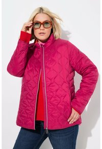 Chaqueta acolchada fucsia con alto cuello, cremallera frontal y forro en contraste azul marino. Combinada con un suéter rojo y jeans de mezclilla.