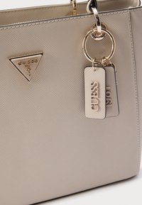 Beige synthetische Tragetasche mit einem Dreieck-Logo, Beschlägen in Goldoptik und zwei Anhängern mit geprägt dargestelltem Text. Strukturierte Oberflächendetails sichtbar.