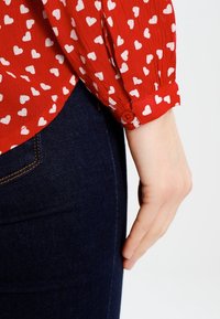 Blouse rouge avec imprimé cœur blanc, tissu texturé, poignet à boutons, associée à un denim bleu foncé. Gros plan sur la manche et la main.