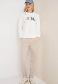 Felpa bianca con cappuccio e stampa colorata "NATURAL", pantaloni jogger beige con strisce laterali e berretto azzurro; abbinati a sneaker beige e blu.
