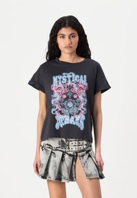 T-shirt en coton noir avec un design graphique coloré portant la phrase "RÊVERIES MYSTIQUES." Associé à une jupe en denim gris plissée.
