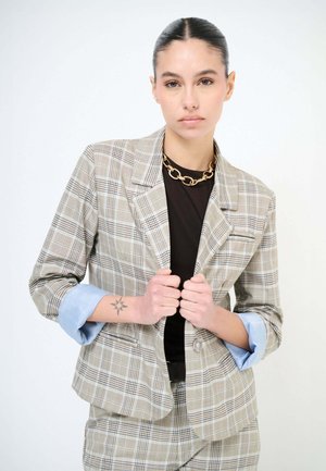 Donna con capelli tirati all'indietro che indossa un blazer a quadri beige e pantaloni abbinati, camicia nera, collana a catena d'oro e con un tatuaggio a forma di stella sul polso.