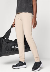 Pantalons de sport beige avec une coupe ajustée et une texture lisse, associés à des baskets noires et un sac de sport noir.