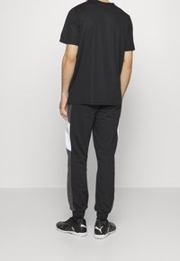 Camiseta negra de cuello redondo, combinada con pantalones de chándal negros y grises con acentos blancos en los laterales. Zapatillas deportivas negras completan el conjunto.