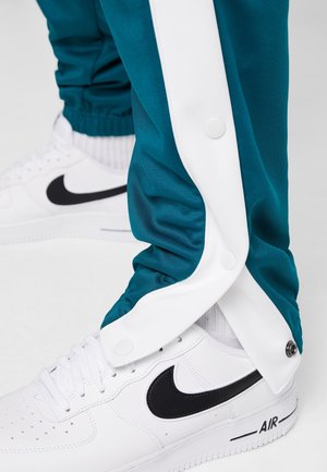 Białe sneakersy Nike Air z czarnym logo, noszone z turkusowo-białymi spodenkami dresowymi na zatrzaski i białymi skarpetkami.