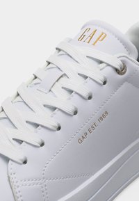 Gros plan d'une basket blanche avec des lacets blancs et la marque "GAP" en or sur la languette, ainsi que l'inscription "GAP EST. 1969" sur le côté.