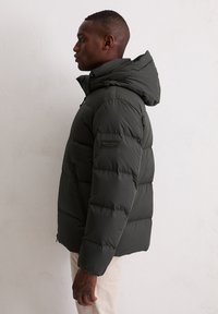 Kurtka puchowa w ciemnozielonym kolorze, z kapturem, teksturowanymi pikowaniami i kieszeniami na zamek. Etykieta Marc O'Polo na lewym rękawie.