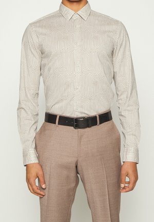 Homme portant une chemise beige à motifs à manches longues, rentrée dans un pantalon marron clair avec une ceinture noire, debout avec les mains détendues.