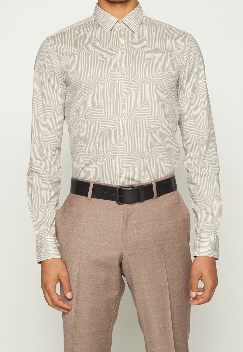 Homme portant une chemise beige à motifs à manches longues, rentrée dans un pantalon marron clair avec une ceinture noire, debout avec les mains détendues.