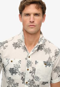 Camisa de manga corta, en un color claro, con un estampado floral en negro. Presenta dos bolsillos en el pecho y botones marrones. Confeccionada en una tela ligera.