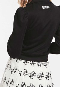 Giacca nera aderente con maniche a sbuffo e una patch con logo sul retro, abbinata a una gonna bianca plissettata con un motivo nero.