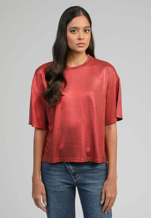 Jeune femme aux longs cheveux noirs portant un haut rouge brillant à manches courtes et amples, ainsi qu'un jean bleu, debout devant un fond gris uni.