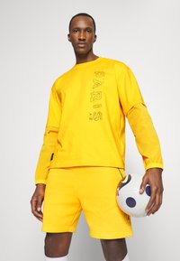 Gul sportssweater med "PARIS" trykt lodret. Sammenstylet med matchende gule shorts. Holder en fodbold. Materiale af bomuldsblanding.