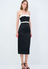 Abito nero senza spalline con una parte superiore bianca a forma di conchiglia, silhouette aderente e una cintura bianca. Modella abbinata a tacchi neri con cinturini.