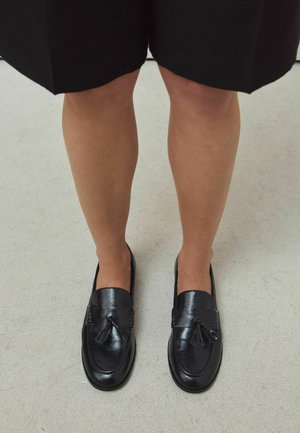 Person wearing black leather tassel loafers and black knee-length skirt standing on a light gray floor.