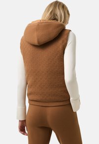 Brun strikket vest med hætte, der har et tekstureret rutemønster og ribbestrikket kant. Lange hvide ærmer er synlige under.
