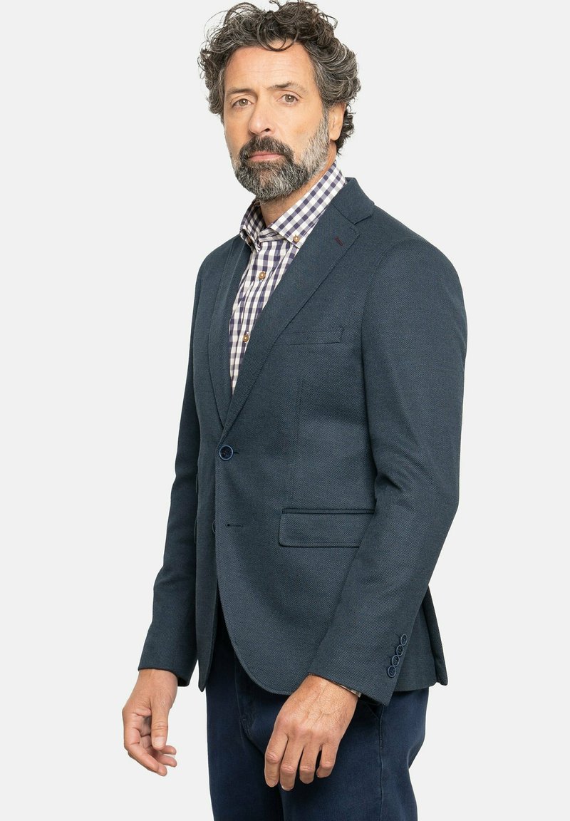 Blazer azul marino con un tejido texturizado, solapas en pico, dos bolsillos frontales y cierre de un solo botón, usado sobre una camisa a cuadros.