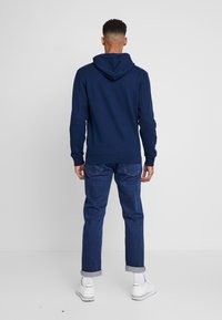 Felpa navy con cappuccio e tasca a marsupio, indossata con jeans in denim scuro con risvolto e sneakers bianche, vista da dietro.