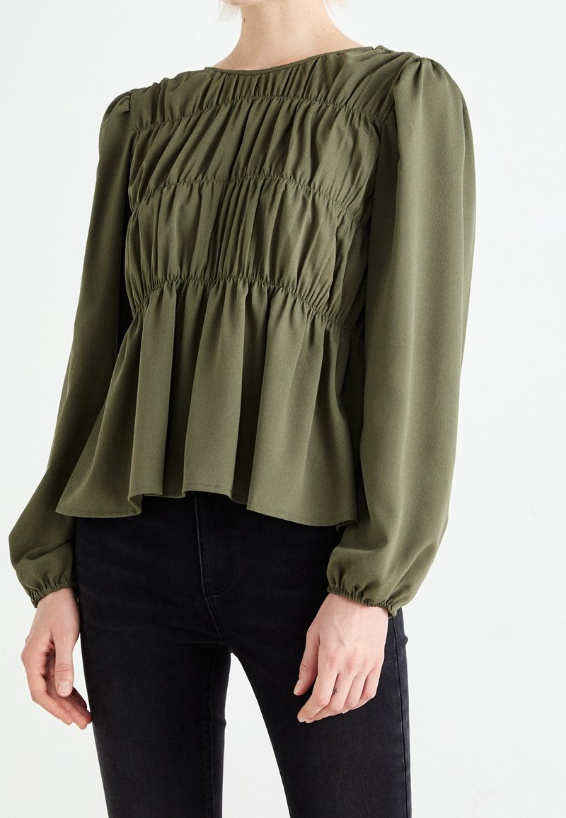 Blouse vert olive à manches longues avec détails de tissu froncé et ourlet évasé. Texture lisse et poignets élastiques, associée à un pantalon noir.