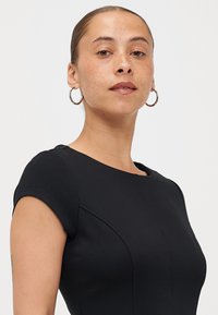 Svart tettsittende kjole med korte ermer, rund halslinje og strukturerte sømmer. Modellen har på seg hoops-øreringer. Glatt tekstur.