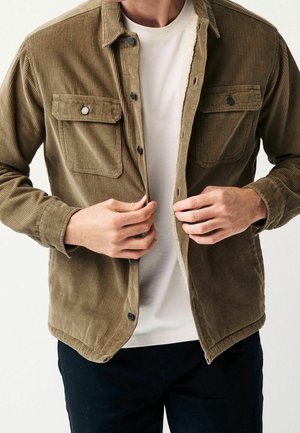 Homme boutonnant une veste en velours côtelé marron avec des poches poitrine, portée sur une chemise blanche et un pantalon foncé.