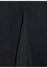 Dettaglio di jeans in denim nero con una cucitura verticale e una piccola fessura, realizzati in tessuto textured. Il design è minimale, senza hardware visibile.