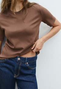 Brun bomullst-shirt med korta ärmar, kombinerad med mörkblå denimjeans med kopparfärgade detaljer och kontrasterande sömmar.