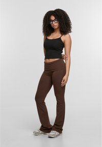 Brown flared leggings made of soft material, paired with a black ribbed tank top. The model wears gray sneakers and large glasses.