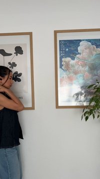 Das Kunstwerk besteht aus zwei gerahmten Drucken mit Holzrahmen. Links: schwarzes florales Grafikdesign. Rechts: bunte Wolkenszene mit Text oben. Eine grüne Pflanze ist sichtbar.
