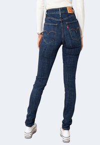 Persona vistiendo jeans ajustados de cintura alta azul oscuro con el logo de Levi's, una camiseta blanca de manga larga, zapatillas Converse blancas, de pie mirando hacia atrás.