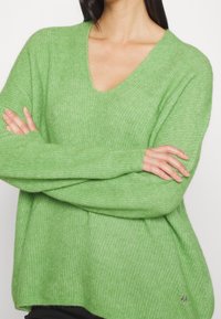 Pull vert texturé avec col en V et manches longues. Coupe ample avec une fente latérale et un accent logo à l'ourlet. Matière douce et légère.