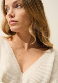 Słoniowa dzianinowa sweter z głębokim dekoltem w kształcie V, prążkowaną teksturą i subtelnym diagonalnym wzorem. Miękki materiał delikatnie opada na ramionach.